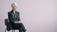 Senior woman with short white hair, Caucasian ethnicity, wearing dark business suit, sitting on chair against purple background, professional portrait for corporate and business use.