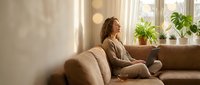 Young woman relaxing on couch with coffee cup in bright living room with plants and warm sunlight streaming through windows creating peaceful atmosphere.