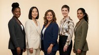 Diverse group of five professional women standing together against neutral background, wearing business casual and formal attire, representing teamwork, leadership and corporate diversity in workplace settings.
