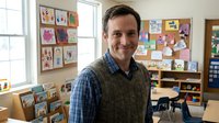 Smiling Caucasian male teacher standing in a colorful elementary school classroom with children artwork on the walls.