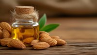Golden almond oil in glass bottle with cork stopper surrounded by raw almonds and green leaves on rustic wooden surface for natural skincare and cooking.