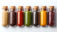 Row of glass spice jars with cork lids containing colorful spices and seasonings including sesame seeds, peppercorns, paprika, herbs and turmeric powder on white background.