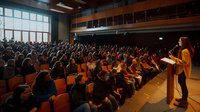 Female speaker addressing large audience in modern auditorium with orange seats. Crowd raising hands during interactive presentation at conference or educational event.