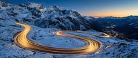 Mountain road with glowing car light trails winding through snowy alpine peaks at dusk, creating golden curves across winter landscape for travel and nature concepts.