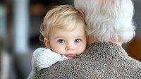 Adorable blonde toddler with blue eyes peeks over grandparent's shoulder while being held, showing innocent curiosity and family bonding in warm indoor setting.
