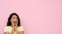 Young woman with dark wavy hair and South Asian ethnicity wearing yellow t-shirt, hands on face expressing surprise and excitement against pink background.