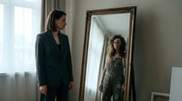 Young woman in black blazer looking at her reflection in ornate wooden mirror frame, contemplating self-image and personal style in modern bedroom interior.