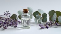 Small glass bottle with cork stopper surrounded by fresh lavender and eucalyptus leaves on white background, perfect for aromatherapy and natural cosmetics concepts.