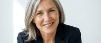 Mature woman with gray hair wearing dark blazer, smiling at camera against white background. Corporate headshot for business, professional profiles, leadership representation.