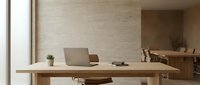 Minimalist wooden desk with laptop and small potted plant in bright, modern office space with natural light and neutral beige walls.