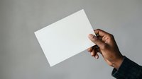 Close-up of male hand with dark skin tone holding blank white card or paper against neutral gray background for mockup and branding purposes.