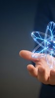 Hand holding glowing neon atom model with blue and cyan energy lines against dark background. Represents nuclear science, physics research, innovation, and technology concepts.