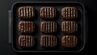 Grilled beef patties with classic sear marks arranged in rows on black plastic tray against dark background, showcasing juicy texture and cooking pattern.