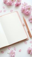 Open pink notebook with rose gold pen surrounded by cherry blossom flowers and petals on white background, feminine flat lay.