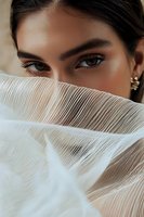 Middle Eastern woman with intense brown eyes and defined brows partially hidden behind sheer pleated white fabric, creating mysterious and elegant portrait.