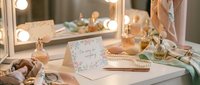 Elegant vanity table with lighted mirror, perfume bottles, pearl jewelry, silk scarves, and a floral apology card in soft pastel tones.