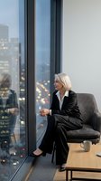 Professional blonde businesswoman in black suit sitting by office window overlooking city skyline at dusk, corporate executive lifestyle concept.