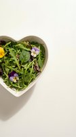 Fresh mixed greens and edible flowers arranged in heart-shaped white bowl on neutral background. Healthy salad with purple and yellow pansy blooms for culinary and wellness content.