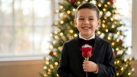 Young boy in formal black suit with red bow tie holding single red rose, smiling at camera with decorated Christmas tree and warm lights in background.