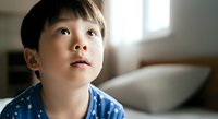 Young Asian boy, approximately 5-8 years old, wearing blue patterned shirt, looking upward with thoughtful expression in bright indoor setting with natural window light and blurred furniture background.