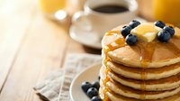 Stack of golden pancakes topped with melting butter and fresh blueberries, served on white plate with coffee and juice for breakfast or brunch.