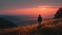 Silhouette of man standing on grassy hill overlooking valley during dramatic orange sunset. Contemplative moment in nature for wellness and travel content.