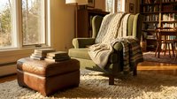 Cozy reading corner with comfortable armchair, knitted throw blanket, stack of books and leather ottoman in warm sunlit home library interior.