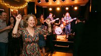 Middle-aged Caucasian woman dancing joyfully at live music venue with band performing on stage. Warm string lights create cozy atmosphere for entertainment.