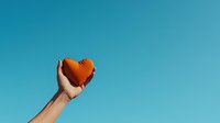 Hand holding orange heart shape against vibrant turquoise sky background, symbolizing hope, love and aspirations in minimalist composition.