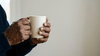 Senior man with dark skin holding white ceramic mug, wearing navy sweater. Hands showing age and texture, isolated against neutral background for beverage, lifestyle and wellness concepts.