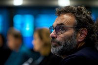 Middle-aged Caucasian man with beard and glasses listening attentively at business conference, with blurred audience and blue background.
