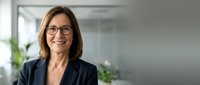 Mature woman with shoulder-length brown hair and glasses wearing dark blazer in modern office environment with plants and natural light, suitable for corporate, business, and professional portraits.