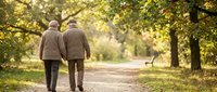 Senior couple walking together on peaceful autumn park path surrounded by golden foliage and warm sunlight creating romantic atmosphere for elderly lifestyle.
