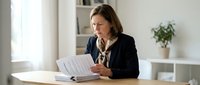 Professional businesswoman reviewing documents at modern office desk with natural lighting and minimalist workspace design for corporate presentations.