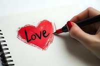Female hand with red manicure writing word love inside sketched heart shape on white notebook page, romantic message concept for valentines day cards.