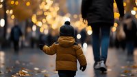 Young child in yellow winter jacket and knit hat walking on city street with bokeh lights and fallen autumn leaves, creating magical evening atmosphere.