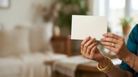 Person with medium skin tone holding blank white card in modern home interior, suitable for greeting cards, invitations, announcements, and promotional messaging.