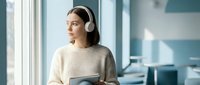 Young woman with medium-length dark hair and light skin tone wearing white over-ear headphones and a beige ribbed sweater, holding a tablet while looking out a window in a modern office environment.