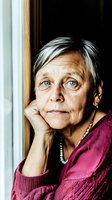 Mature woman with striking blue eyes and silver hair in pink sweater gazes thoughtfully by window, hand resting on chin. Natural lighting creates dramatic portrait.
