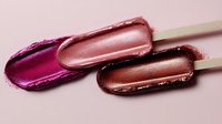 Luxurious metallic lip gloss swatches in vibrant pink and bronze tones on wooden applicators against soft pink background, showcasing rich pigmentation and glossy finish.