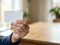 Senior male hand holding blank white card at wooden table with blurred office background, suitable for business messaging, advertising, and corporate communications.