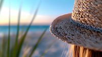 Straw sun hat detail against blurred beach background with sea and sunset, grass blade in foreground creates summer vacation mood and coastal atmosphere.