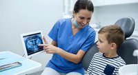 Female dentist in blue scrubs showing dental X-ray results to young Caucasian boy patient in striped shirt at modern dental clinic office.