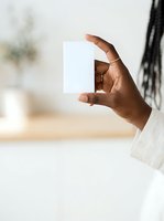 Hand of a dark-skinned man wearing a white linen shirt holding a blank white card or paper mockup against a blurred interior background with natural lighting.