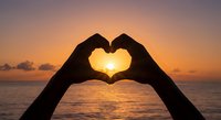 Hands forming heart shape silhouette against golden sunset over ocean water. Romantic gesture symbol of love and affection during magical twilight hour at beach.