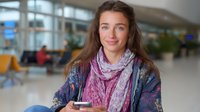 Young Caucasian woman with long brown hair wearing floral jacket and pink scarf smiling while holding smartphone in airport terminal waiting area with blurred passengers.