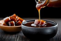 Rich caramel sauce being poured into dark bowl with salted caramel candies on wooden spoon, perfect for dessert recipes and culinary photography.