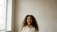 Young woman with curly brown hair and medium skin tone wearing white shirt, smiling against neutral beige wall with natural window light for portrait and lifestyle use.