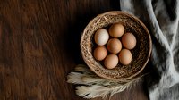 Fresh brown eggs in rustic woven basket with dried wheat stalks and soft fabric on dark wooden background, moody farm still life composition.