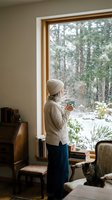 Young woman in cozy sweater and beanie holding warm drink by window watching winter snowfall in comfortable living room with books and furniture.